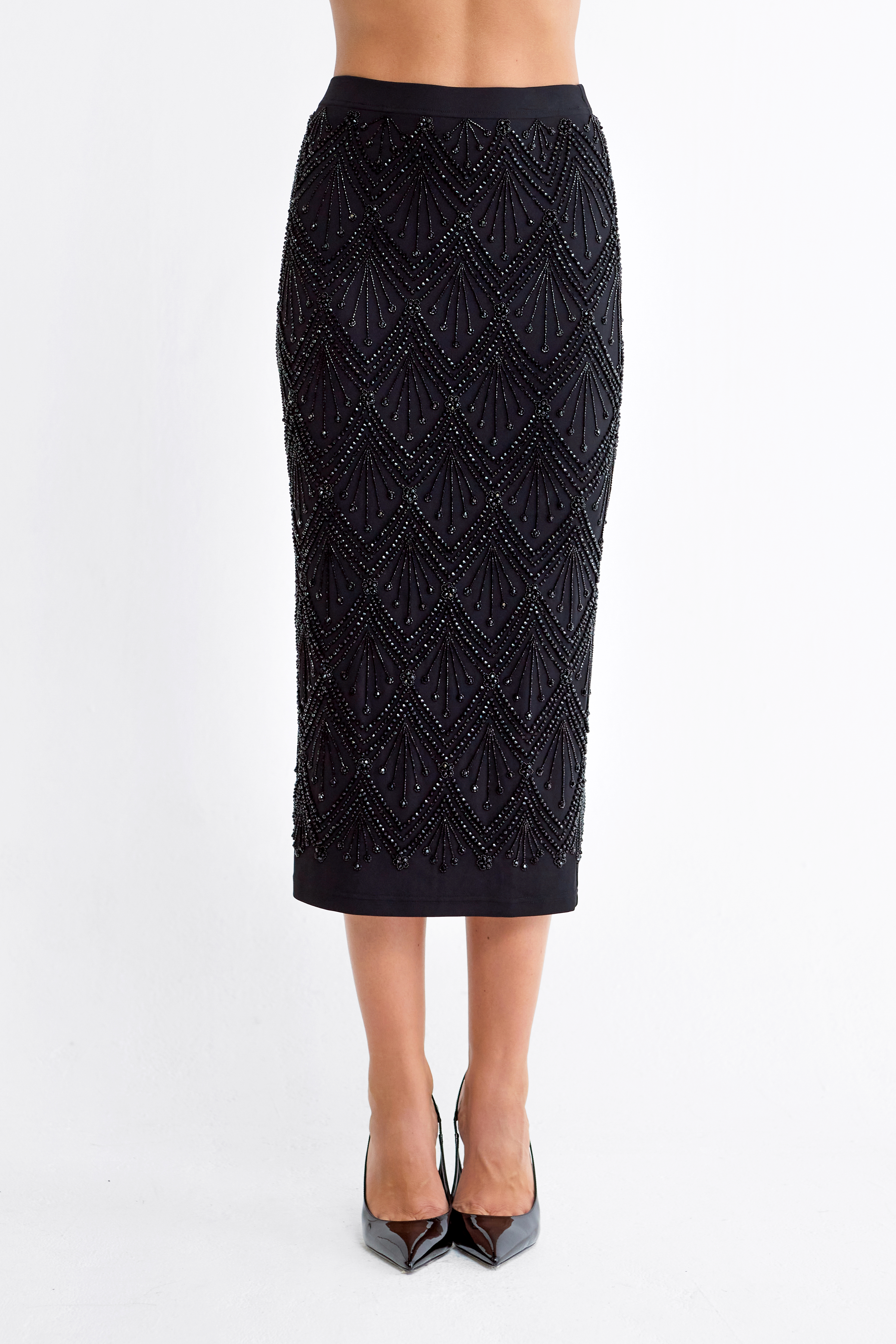 A black midi skirt with an intricate geometric beaded pattern, shown on a model from the waist down wearing black pointed-toe heels against a plain white studio background.