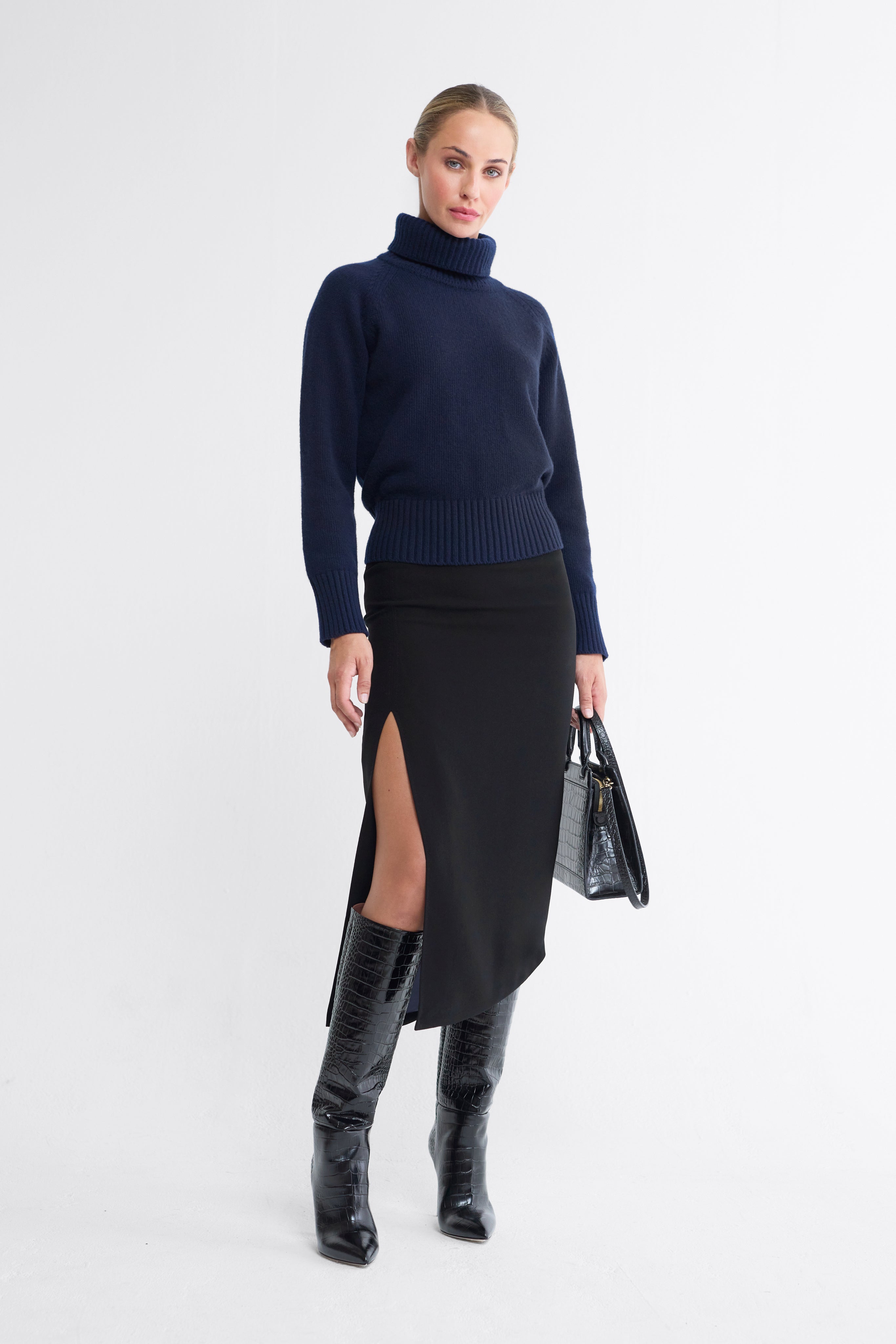 A model wearing a navy blue turtleneck sweater, a black asymmetrical skirt with a thigh-high slit, and black knee-high boots, while holding a black handbag.